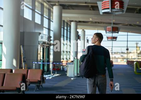 Rückansicht eines nicht erkennbaren männlichen Reisenden, der mit Rucksack am Flughafen entlang läuft. Rückansicht des Mannes, der auf dem Weg zum Flug am Gate des Terminals vorbeikommt Stockfoto