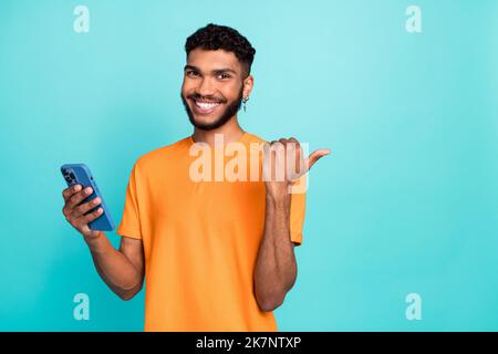 Foto von guter Laune lustigen Kerl tragen orange T-Shirt Chat moderne Gadget Daumen zurück leeren Raum isoliert blaugrün Farbe Hintergrund Stockfoto