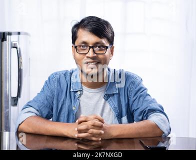 Porträt eines jungen indischen Mannes mittleren Alters, der eine Brille trägt und zu Hause mit einem lächelnden Gesicht auf weißem Hintergrund am Tisch sitzt Stockfoto