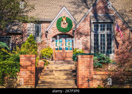 Rustikales gehobenes Ziegelwohnhaus mit Magnolienblütenkranz an Doppeltüren und großem Weihnachtskranz oben - mit amerikanischer Flagge - Nahaufnahme Stockfoto