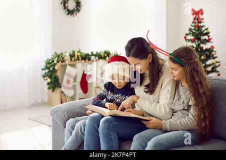 Liebende Mutter mit ihren Kindern lesen Buch der Geschichten in gemütlichen hellen Wohnzimmer an verschneiten Wintertag. Stockfoto