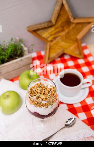 Kirschjoghurt-Parfait mit knusprigem Müsli oben in einem Weinglas und einer Tasse Tee Stockfoto