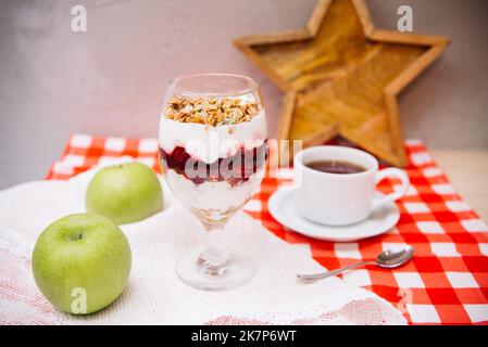 Kirschjoghurt-Parfait mit knusprigem Müsli oben in einem Weinglas und einer Tasse Tee Stockfoto