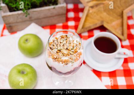 Kirschjoghurt-Parfait mit knusprigem Müsli oben in einem Weinglas und einer Tasse Tee Stockfoto