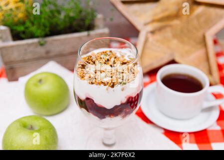 Kirschjoghurt-Parfait mit knusprigem Müsli oben in einem Weinglas und einer Tasse Tee Stockfoto