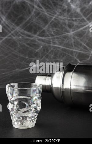 Ein transparentes Glas in Form eines Schädels, im Hintergrund ein Shaker, auf schwarzem Hintergrund mit Spinnennetz. Halloween-Konzept Stockfoto