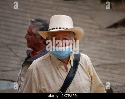 Jericó, Antioquia, Kolumbien - April 5 2022: Der alte Mann, der eine Cyan-Maske, einen gelben Hut und ein Hemd verwendet, schaut auf die Kamera Stockfoto