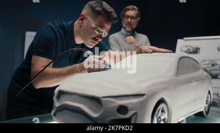 Der Automobilingenieur arbeitet mit einem rotierenden Werkzeug an der Skulptur eines Prototyps eines Automodells, während er mit seinem leitenden Kollegen diskutiert. High-Tech-Automobildesign-Unternehmen mit innovativen Technologien. Stockfoto