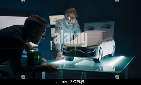 Erfahrene Automobilingenieure diskutieren das Fahrzeugdesign vor einem Prototyp-Modell der Skulptur, das mit einem grünen Laserlicht auf einem Glastisch für Ausrichtung und Perfektion aufgestellt wird. Stockfoto