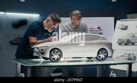 Zwei erfahrene Automobilingenieure diskutieren das Fahrzeugdesign anhand der Skizzen auf dem Brett. Korrekturen an der Prototyp-Rechen-Skulptur mit einem rotierenden Werkzeug mit drehender Spitze. Stockfoto