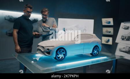 Erfahrene Automobildesigner und -Entwickler diskutieren in einem Hightech-Autorabor vor einem Rake-Modellauto-Prototyp das Design. Nehmen Sie Designkorrekturen mit einem digitalen Touchscreen-Tablet vor. Stockfoto