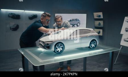 Erfahrene Automobildesigner und Autoentwickler diskutieren das Design vor einem auf einem Glastisch platzierten Rechen-Modell. Ein High-Tech-Laborraum mit LED und Skizzen. Stockfoto