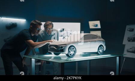 Erfahrene Automobildesigner und Autoentwickler diskutieren das Design vor einem auf einem Glastisch platzierten Rechen-Modell. Ein High-Tech-Laborraum mit LED und Skizzen. Stockfoto