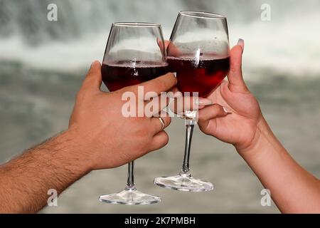 Nahaufnahme, Hände eines Mannes und einer Frau, die mit einem Glas Wein auf einem verschwommenen Wasserfall-Hintergrund toasten. Stockfoto