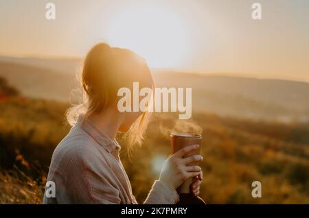 Frau genießt heißen Tee aus der Thermoschale bei Sonnenuntergang auf einem Hügel mit ländlichem Hintergrund Stockfoto