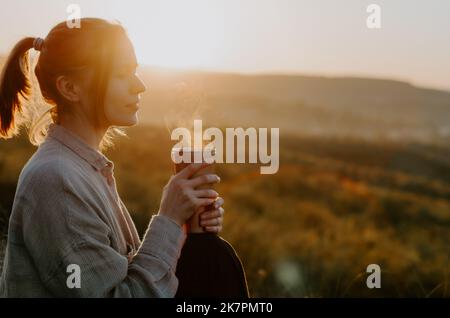 Frau genießt heißen Tee aus der Thermoschale bei Sonnenuntergang auf einem Hügel mit ländlichem Hintergrund Stockfoto