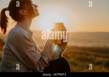 Frau genießt heißen Tee aus der Thermoschale bei Sonnenuntergang auf einem Hügel mit ländlichem Hintergrund Stockfoto