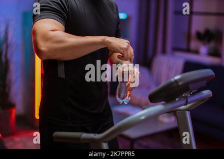 Nahaufnahme der Hände des Sportmanns trinken Öffnung Wasserflasche, tun Fitness-Übung, Laufen auf dem Laufband . Athletischer und muskulöser Mann, der in der modernen Wohnung nachts trainieren kann. Stockfoto
