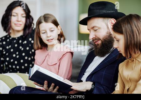 Seitenansicht Porträt eines orthodoxen jüdischen Mannes, der im häuslichen Umfeld ein Buch für Kinder liest Stockfoto