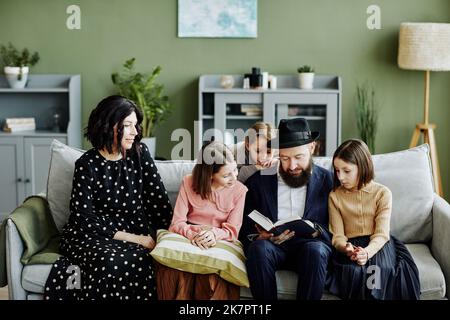 Porträt einer jüdischen Familie mit drei Kindern, die gemeinsam Buch lesen, in einem modernen Zuhause Stockfoto