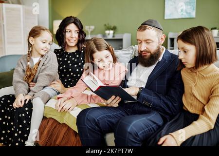 Porträt eines orthodoxen jüdischen Mannes, der Kippah trug, während er viele Kinder zu Hause beim Lesen des Buches vortrug Stockfoto