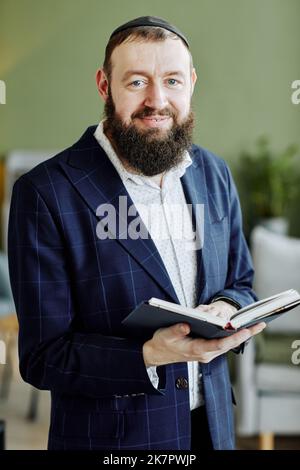 Vertikales Porträt eines bärtigen jüdischen Mannes mit Kippah und Blick auf die Kamera, die Buch hält Stockfoto