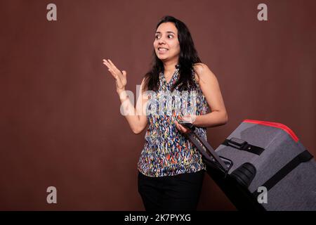 Fröhlicher indischer Reisender, der mit fahrbaren Koffern stand, sprach, in den Urlaub ging. Frau mit rollendem Gepäck, lächelnder Tourist, der Gepäck am Flughafen trägt, Tourismuskonzept Stockfoto