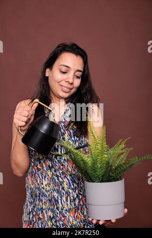 Lächelnde indische Frau, die Topfpflanze wässert, wachsende Laubpflanze. Lady hält kann, Pflege über exotische Indoor-Blumentopf, Person, die auf tropische grüne Blätter, Hobby-Lifestyle Stockfoto