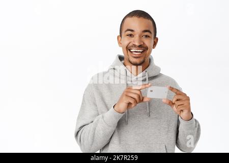 Porträt des glücklichen jungen Mannes erhält seine Bankkarte, zeigt Kreditkarte und lächelt, stehend auf weißem Hintergrund Stockfoto