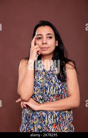 Nachdenkliche indische Frau schaut seitwärts Porträt, Modell posiert, hält Finger auf dem Gesicht. Nachdenkliche Person, die mit zweifelhaftem Gesichtsausdruck steht, Vorderansicht Studio, mittlere Aufnahme Stockfoto