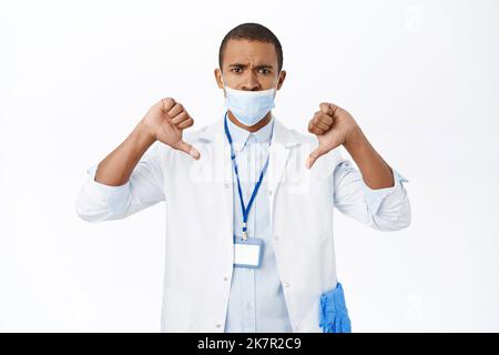 Arzt, der während der Covid-19-Pandemie mit falschen medizinischen Masken verärgert ist, zeigt Daumen nach unten, steht auf weißem Hintergrund Stockfoto