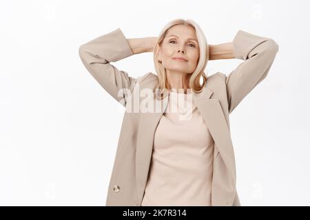 Porträt einer glücklichen Frau mittleren Alters, einer schönen älteren Dame, die sich ausruhte, die Hände hinter dem Kopf hielt und sich entspannte, zufrieden lächelte, über Weiß stehend Stockfoto