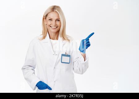 Lächelnder Arzt in medizinischen Handschuhen zeigt nach rechts, zeigt Werbung, Klinik-Logo, steht auf weißem Hintergrund Stockfoto
