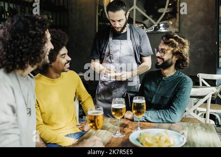 Junge Kellnerin nimmt Bestellung von multirassischen männlichen Freunden im Restaurant Stockfoto