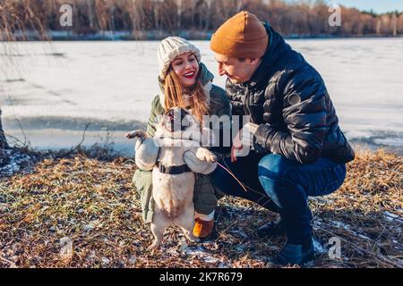Glückliches junges Paar, das im verschneiten Winterpark einen Mops-Hund spaziert. Menschen entspannen sich beim Spielen und Spaß mit Haustieren im Freien. Aktivitäten mit Tieren Stockfoto