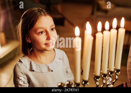 Hochwinkelporträt eines lächelnden jüdischen Mädchens, das während der Chanukka-Feier die Menorah-Kerze ansah Stockfoto