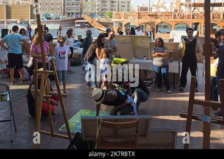 Vielfältiges Publikum und Medien auf einer Kunstmesse im Freien in Dubai sehen die VAE wie eine Künstlerin vor Ort eine Leinwand malt. Ein männlicher Videofilmer nimmt eine Nahaufnahme auf. Stockfoto