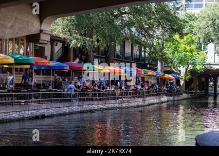 Auf dem River Walk entlang des San Antonio River in der Innenstadt von San Antonio, Texas, sitzen die Menschen an Tischen, die mit bunten Sonnenschirmen bedeckt sind. Am 25.. September 20 Stockfoto