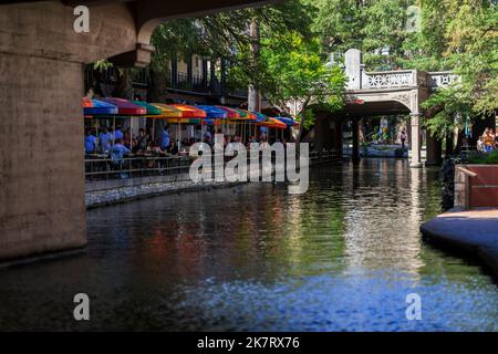 Auf dem River Walk entlang des San Antonio River in der Innenstadt von San Antonio, Texas, sitzen die Menschen an Tischen, die mit bunten Sonnenschirmen bedeckt sind. Am 25.. September 20 Stockfoto