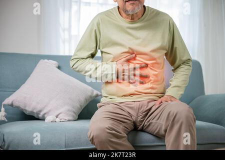 Älterer asiatischer Mann, der auf dem Sofa sitzt, an Magenschmerzen leidet und seine Bauchschmerzen hält, unglücklicher alter Mann, der sich wegen des Essens unwohl fühlt Stockfoto