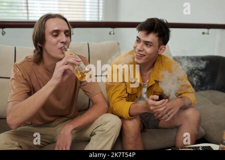 Fröhliche junge Menschen, die vap rauchen und Bier trinken, wenn sie sich zu Hause entspannen Stockfoto