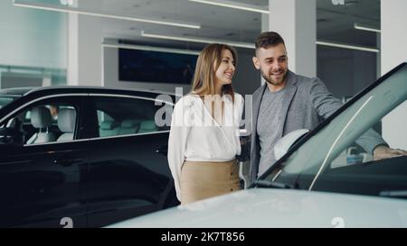 Attraktive junge Paar zuversichtlich bärtigen Kerl und seine lächelnde hübsche Freundin sind die Wahl neuer Automobil zusammen Blick auf Luxus-Auto in Autohaus und sprechen. Stockfoto