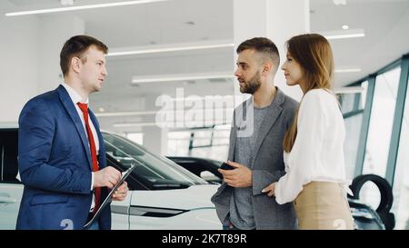Interessierte junge Paar Mann und Frau sprechen mit Manager im Autohaus, während er ihnen über Luxus-Auto-Modell erzählt. Schöne Menschen, Transport und Geschäftskonzept. Stockfoto