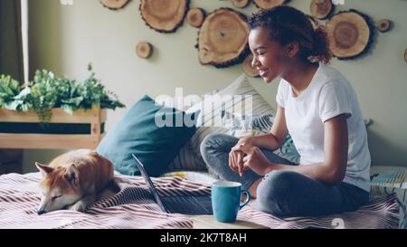 Glückliches Mädchen ist skyping im Gespräch online mit Freunden gestikulieren und Blick auf Laptop-Bildschirm sitzen auf dem Bett zu Hause mit niedlichen Welpen liegen neben ihr. Kommunikations- und Technologiekonzept. Stockfoto