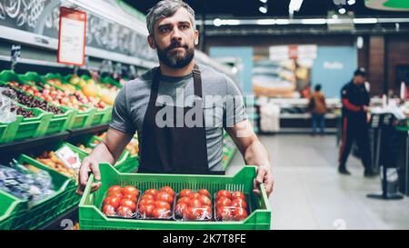 Junger bärtiger Verkäufer in der Schürze, der eine Schachtel Tomaten im Supermarkt in der Obst- und Gemüseabteilung trug. Verkäufer, Lebensmittelgeschäft und Prosezession Konzept. Stockfoto