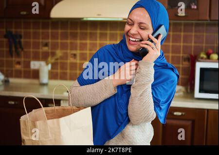 Entzückende Frau aus dem Nahen Osten in Hijab, lacht am Telefon, während sie Einkaufstasche mit Lebensmitteln in der Küche auspacken Stockfoto