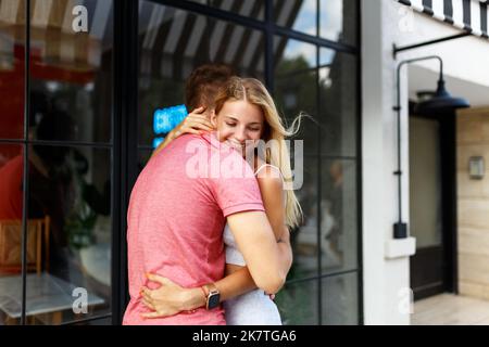 Abschiedskonzept. Erfreut glücklich lächelnde blonde Frau sagt Auf Wiedersehen oder hallo zu Freund, gibt warme Umarmung, Pose zusammen gegen Café-Platz. Wahrheitsgemäße Gefühle Stockfoto