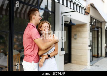 Abschiedskonzept. Erfreut glücklich lächelnde blonde Frau sagt Auf Wiedersehen oder hallo zu Freund, gibt warme Umarmung, Pose zusammen gegen Café-Platz. Wahrheitsgemäße Gefühle Stockfoto