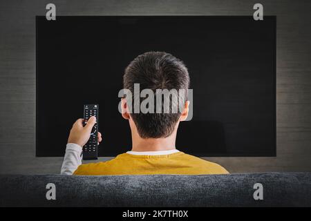 Mann, der Fernsehen guckend, den Plasma-Flachbildfernseher anschminkend, die Fernbedienung auf einen leeren Fernsehbildschirm an der Wand gerichtet. Mann wechselt zu Hause, zurück, die Kanäle Stockfoto