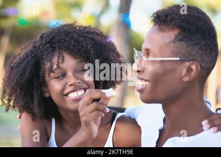 Fröhliche Frau lächelt und hat Spaß, ihren Freund zu füttern, während sie gemeinsam ein Picknick im Freien in einem Park genießt. Stockfoto